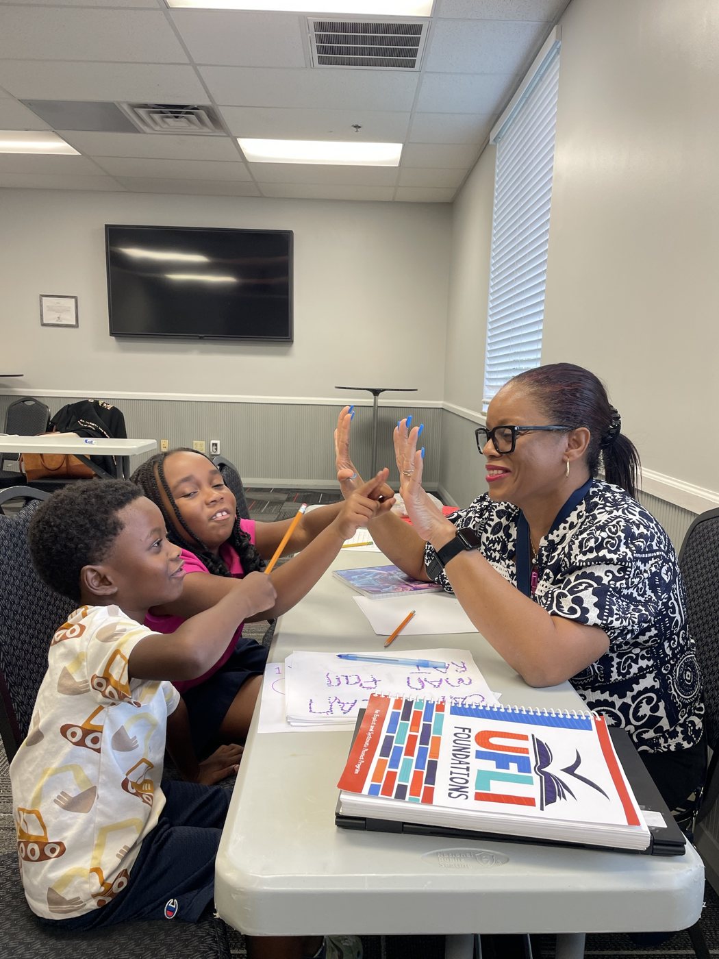 A tutor smiles and does a hand-motion activity with two young students at a table, with a UFLI Foundations phonics manual and word practice sheets in front of them.